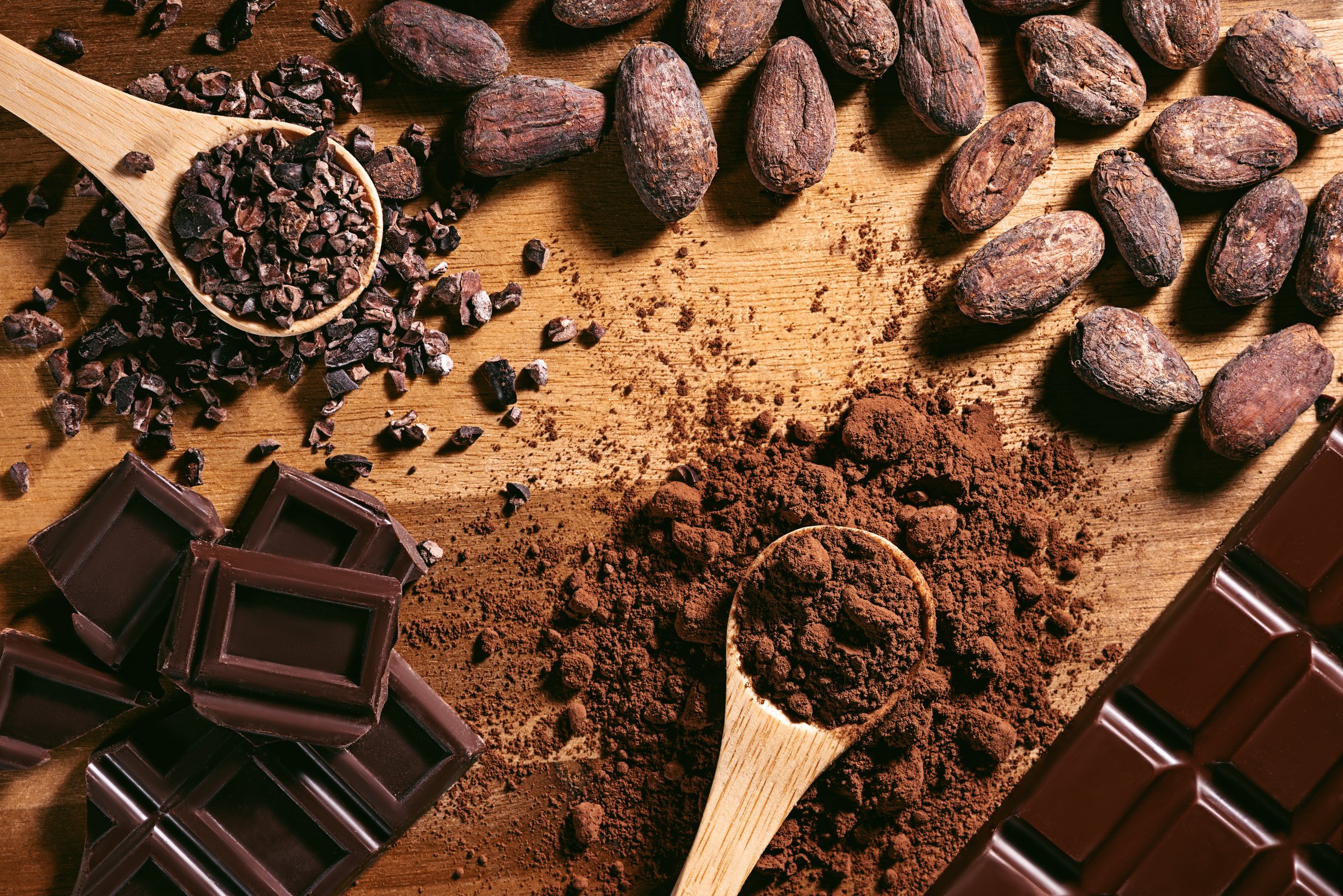 Chocolate bar, cocoa powder, cacao beans and nibs, heap in wooden spoons, chocolate background